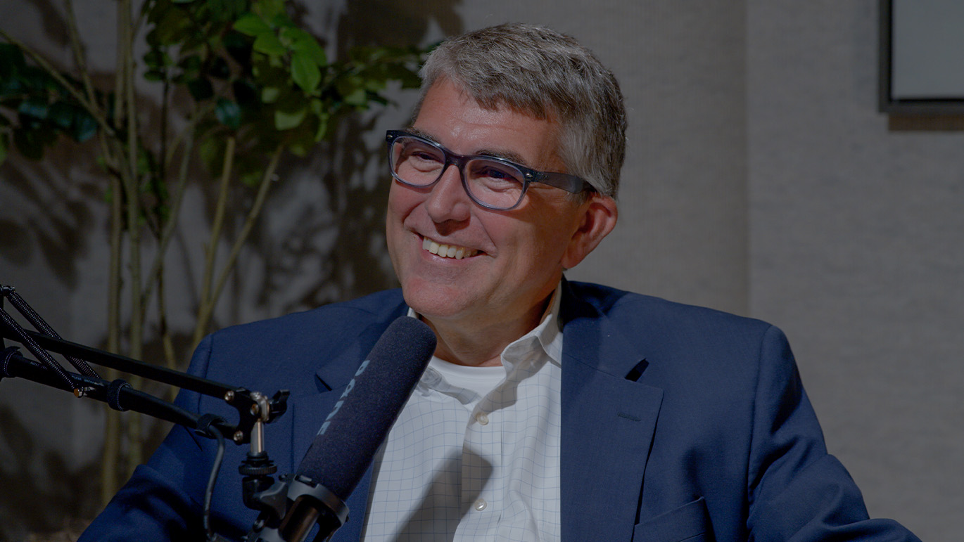 Person seated in a podcast studio speaking into a microphone while wearing glasses, a white button-down shirt, and a blue blazer.