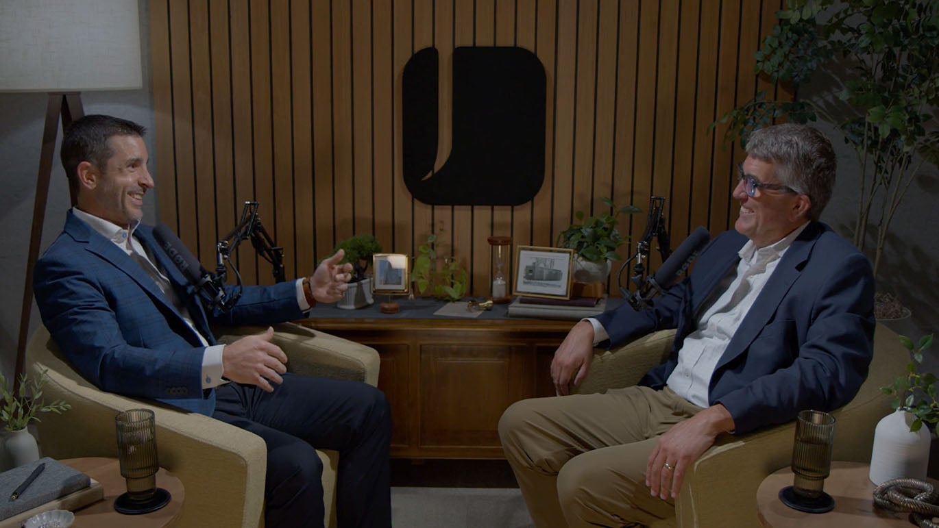 Two podcast hosts sit across from each other in armchairs, speaking into microphones in a studio with wood-paneled walls, plants, and a central table.