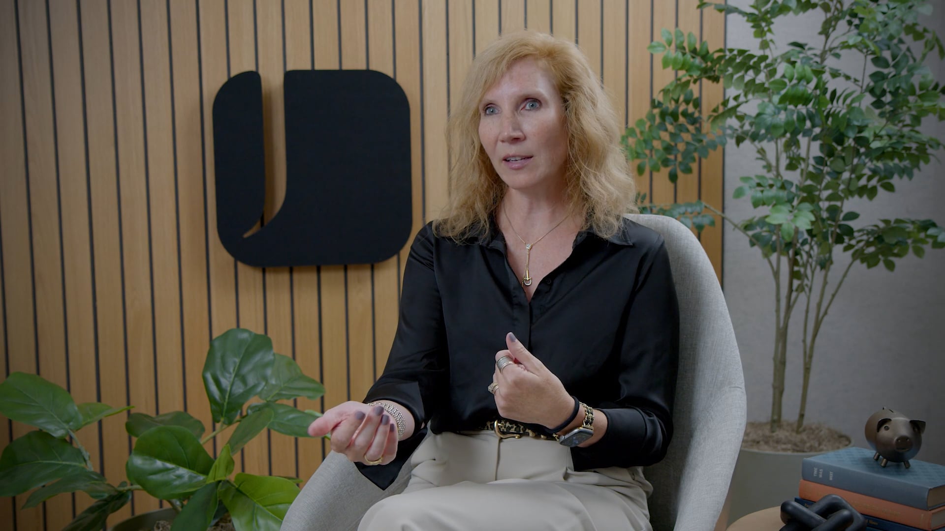 Anita wearing a black blouse and light-colored pants seated in a modern office setting, gesturing while speaking, with plants and wood-panel wall accents behind them.