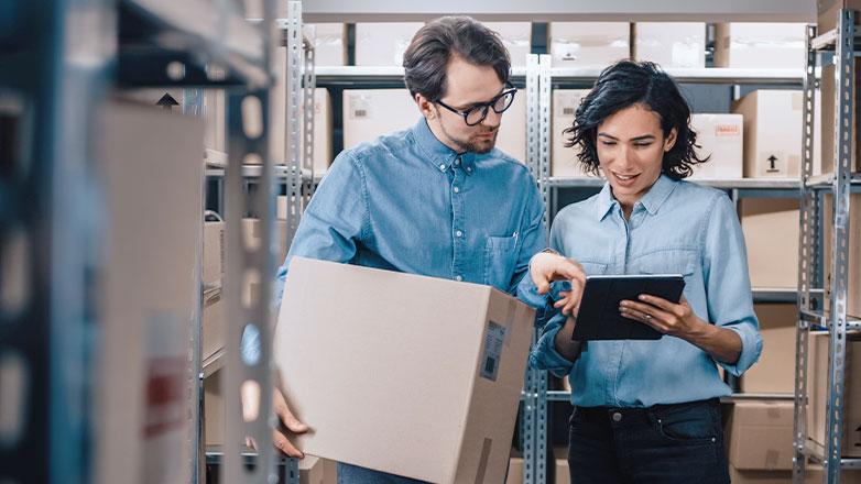 man holding box in warehouse pointing at tablet held by woman