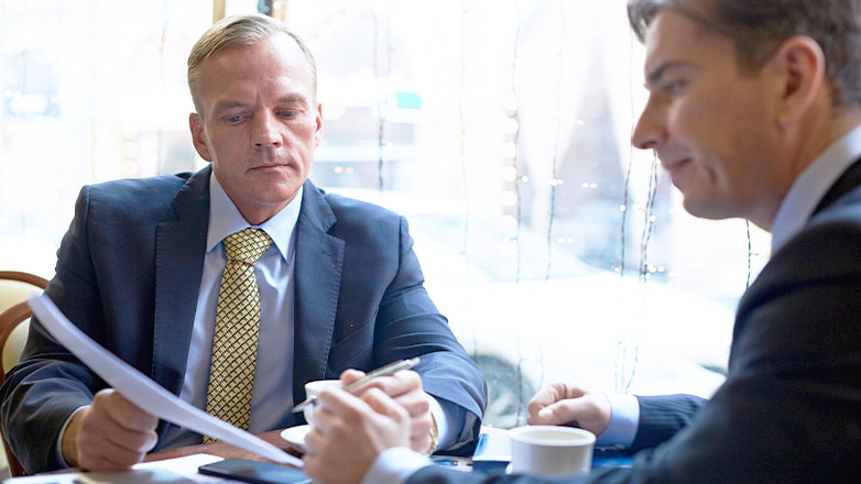 two men meeting at table by window discussing papers in hand