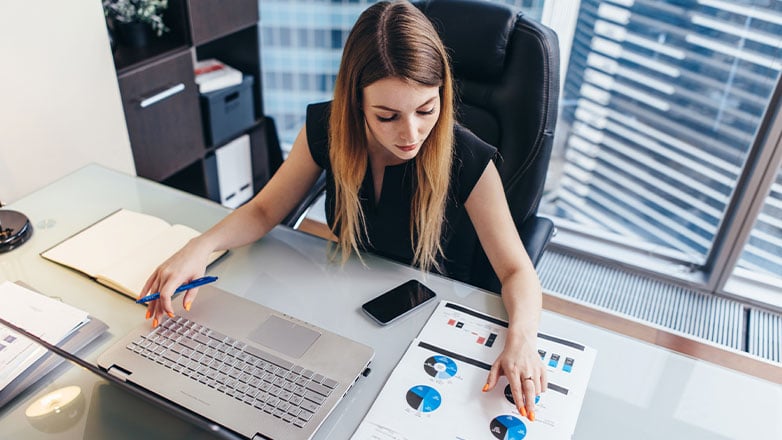 woman at desk comparing numbers on paper to laptop