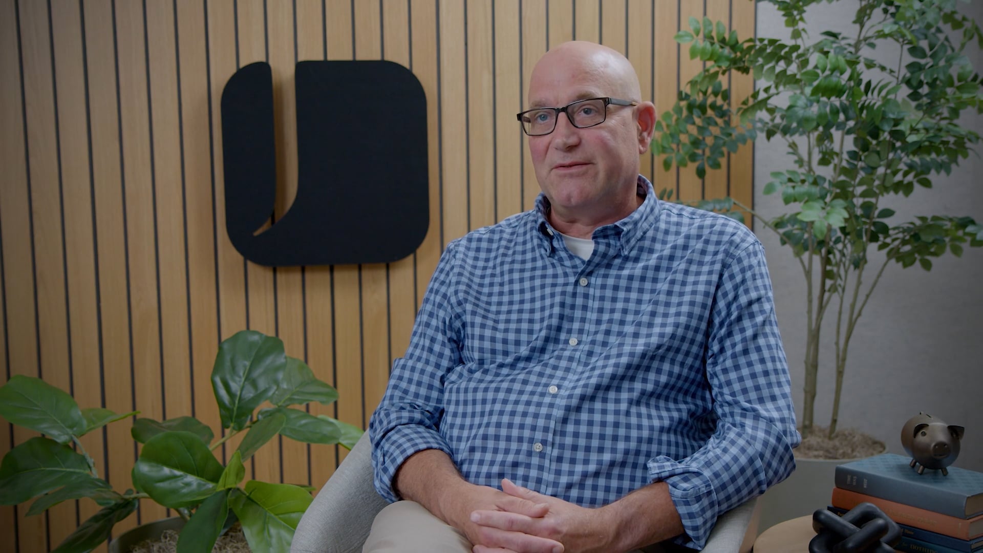 John Keller seated in a chair during an office interview, with wood-slat wall paneling, indoor plants, and a company logo in the background.