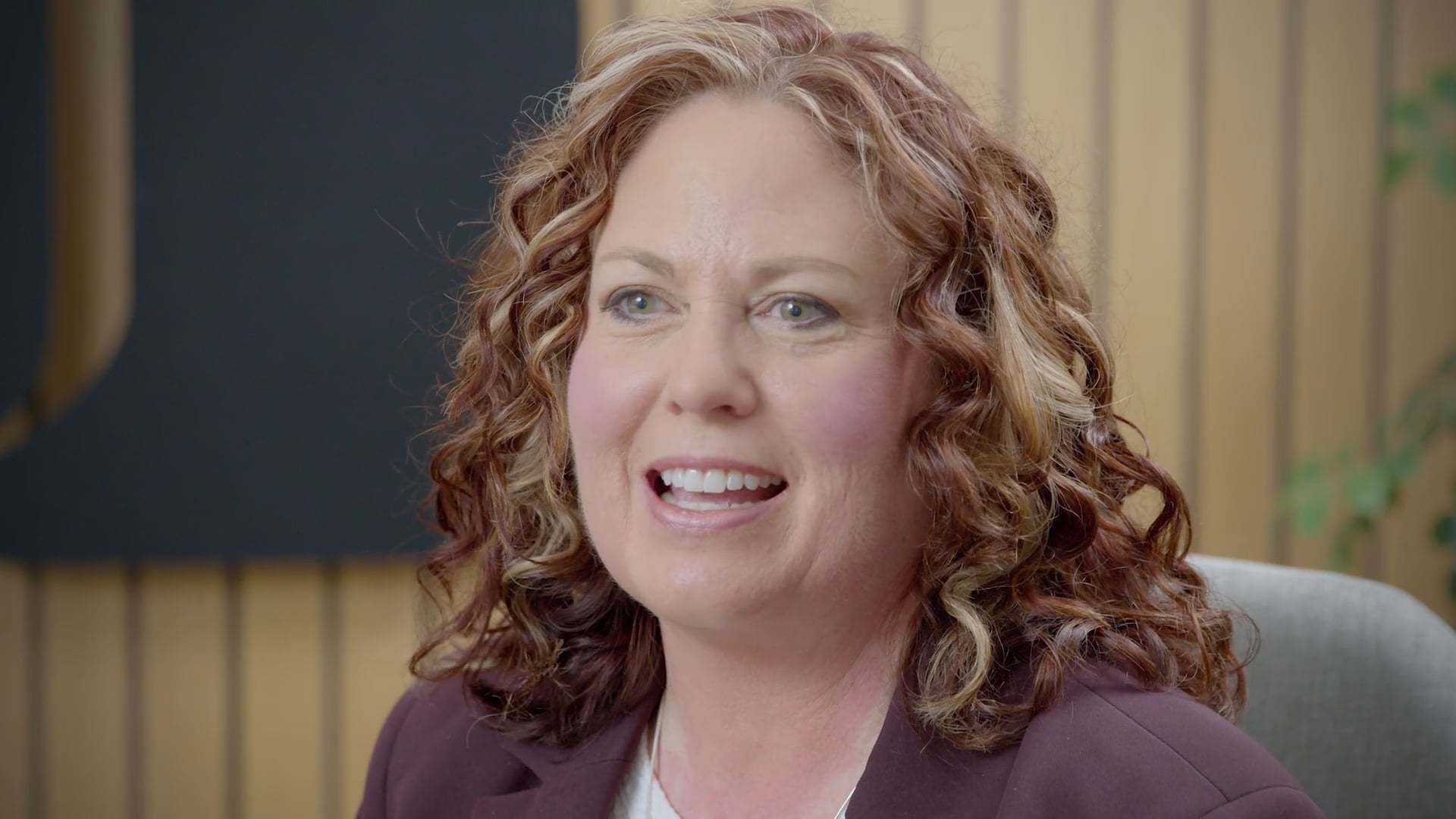 Close-up portrait of a person with curly hair seated indoors, wearing a dark blazer and light-colored top. The background features warm wood paneling and soft office furnishings, creating a professional setting.