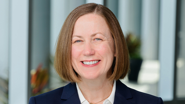 Professional headshot of a person with short, straight hair wearing a navy blazer and white shirt, smiling at the camera, with a softly blurred office background.