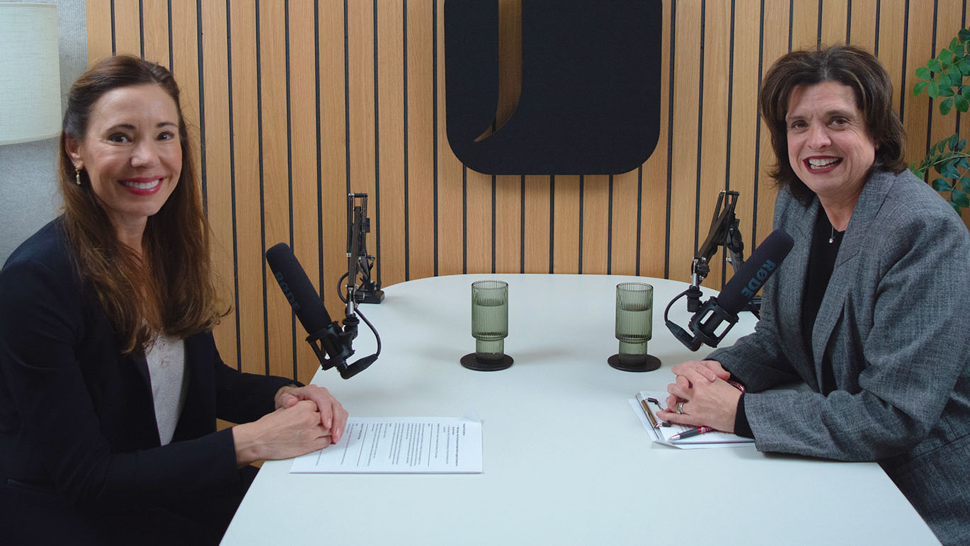 Two women sitting in a podcast studio smiling at the camera.