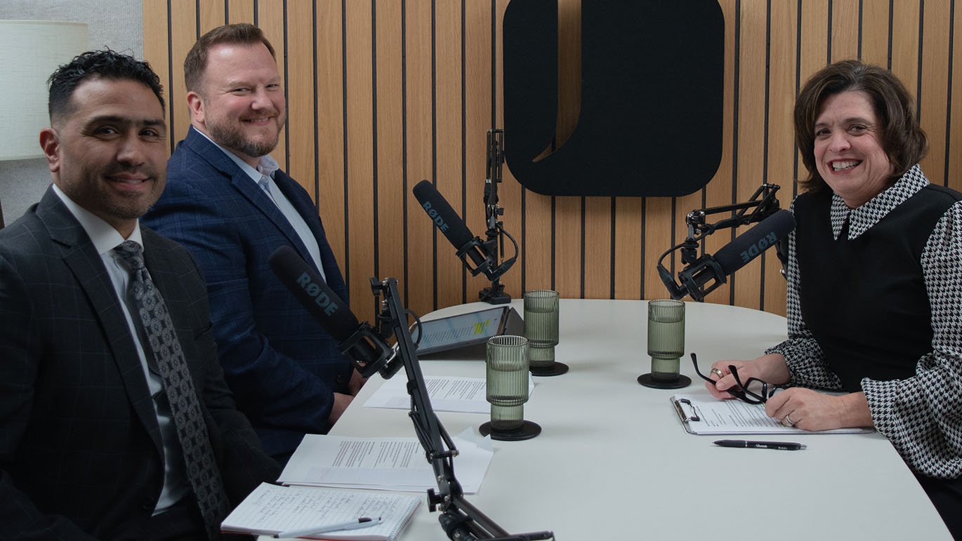 Two men and a woman sit at a white table in a recording studio, smiling at the camera, with three microphones on boom arms in front of them.