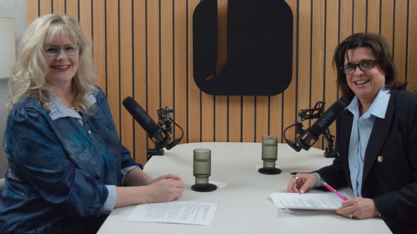 Two individuals seated at a round table in a recording studio with microphones, holding printed notes, against a wood-paneled wall with a large logo.