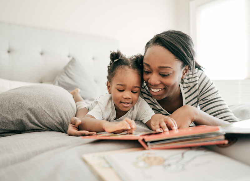 Mom reads her toddler daughter a book while they lay on the bed.