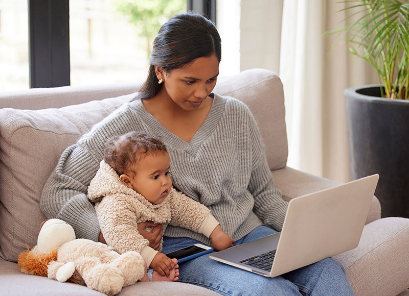 Mom cuddles with baby on a couch while working on a laptop.