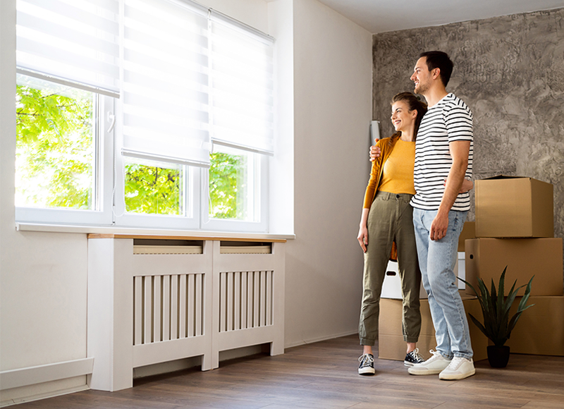 A young couple is standing in a new house, looking out the window. The woman is smiling and the man has his arm around her shoulder. There are boxes and a plant next to them.