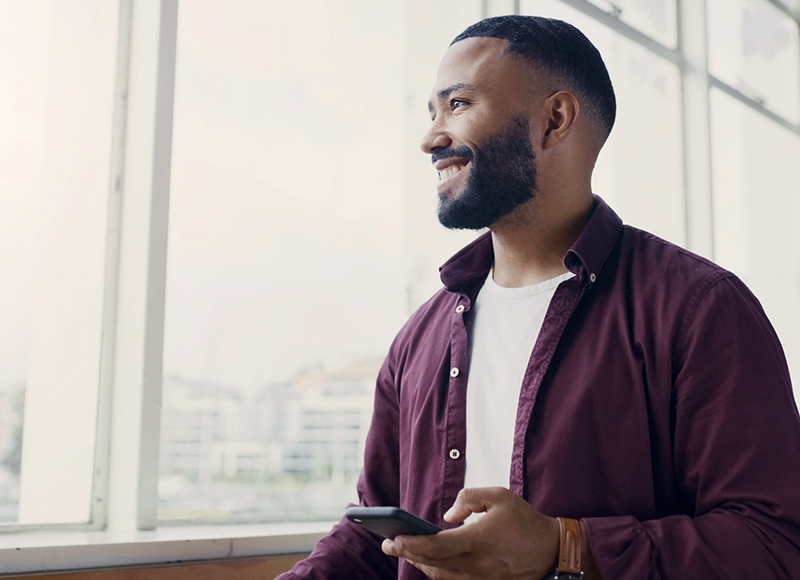 A young African-American man is standing in front of a window. He is wearing a maroon button-down shirt and has a beard. He is smiling and looking to the left. He is holding a phone in his right hand.