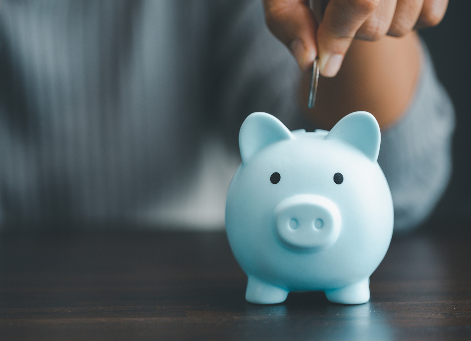 A hand releases a coin into a light blue piggy bank with a slot on top.