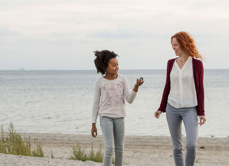 Mom and daughter walking barefoot together on a sandy beach.