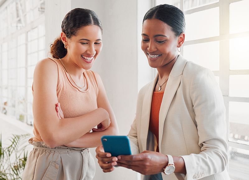 Two young women are looking at something on a smartphone together. They are both smiling and appear to be enjoying themselves.