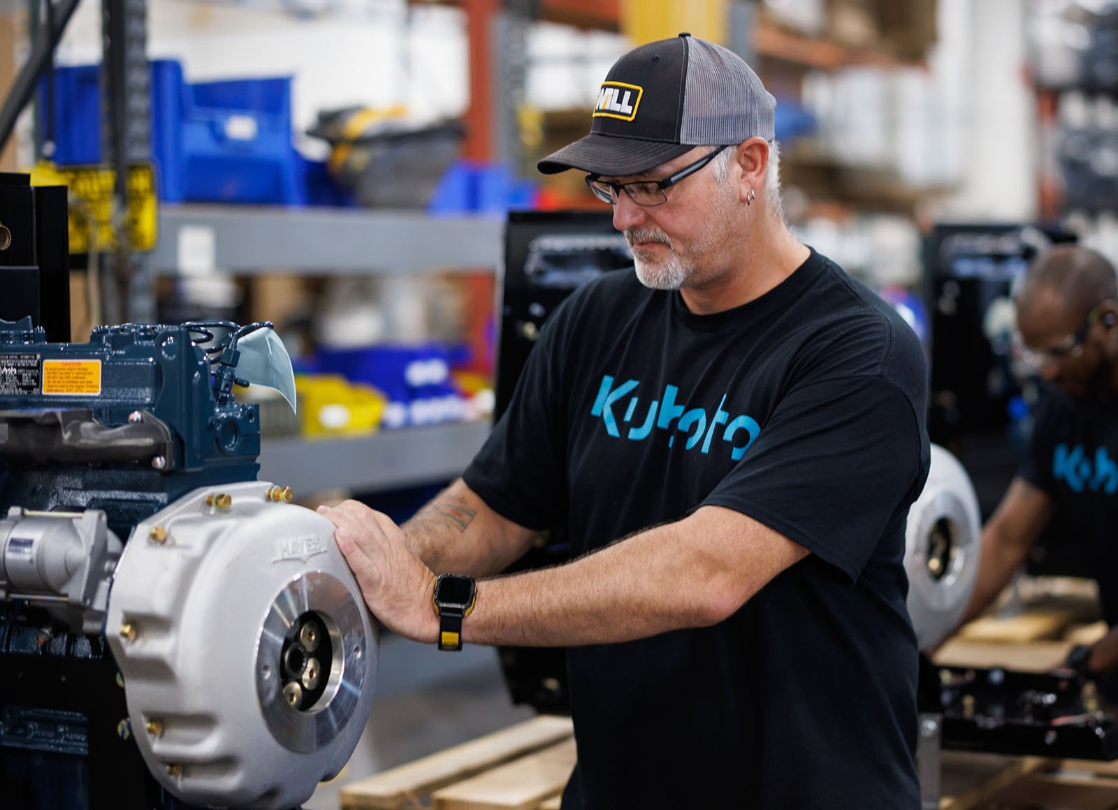 A man wearing a Kubota shirt is crouched over, performing maintenance on a engine.